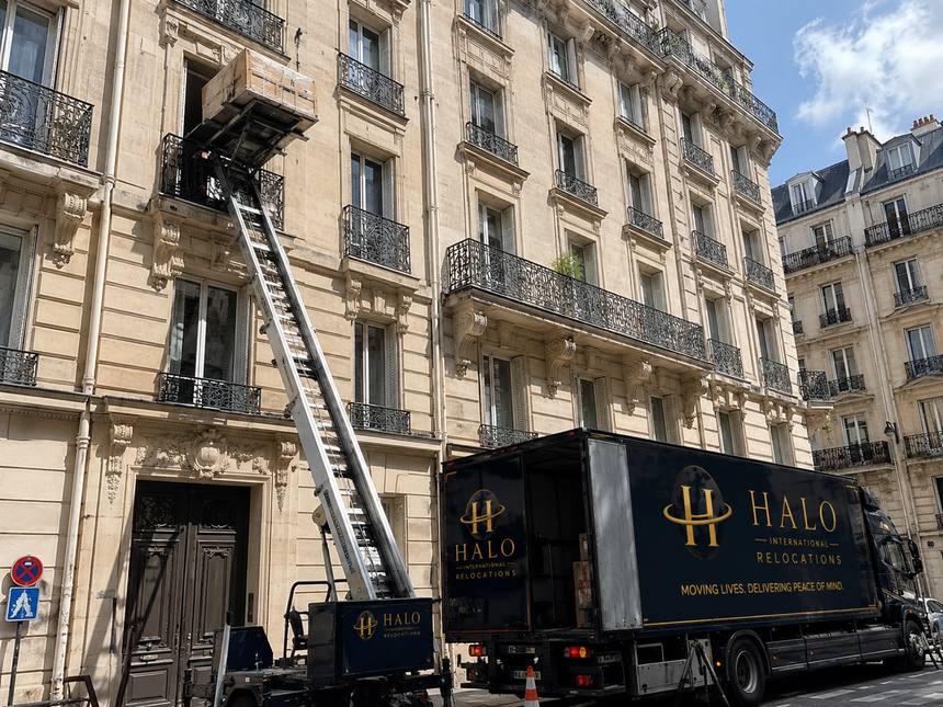 A furniture lift raised to a fourth-floor window on a Paris street, with a Halo lorry parked below